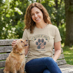 Woman in a tan "Rescued by Grace Paws & Prayers" t-shirt petting a small dog on a park bench.