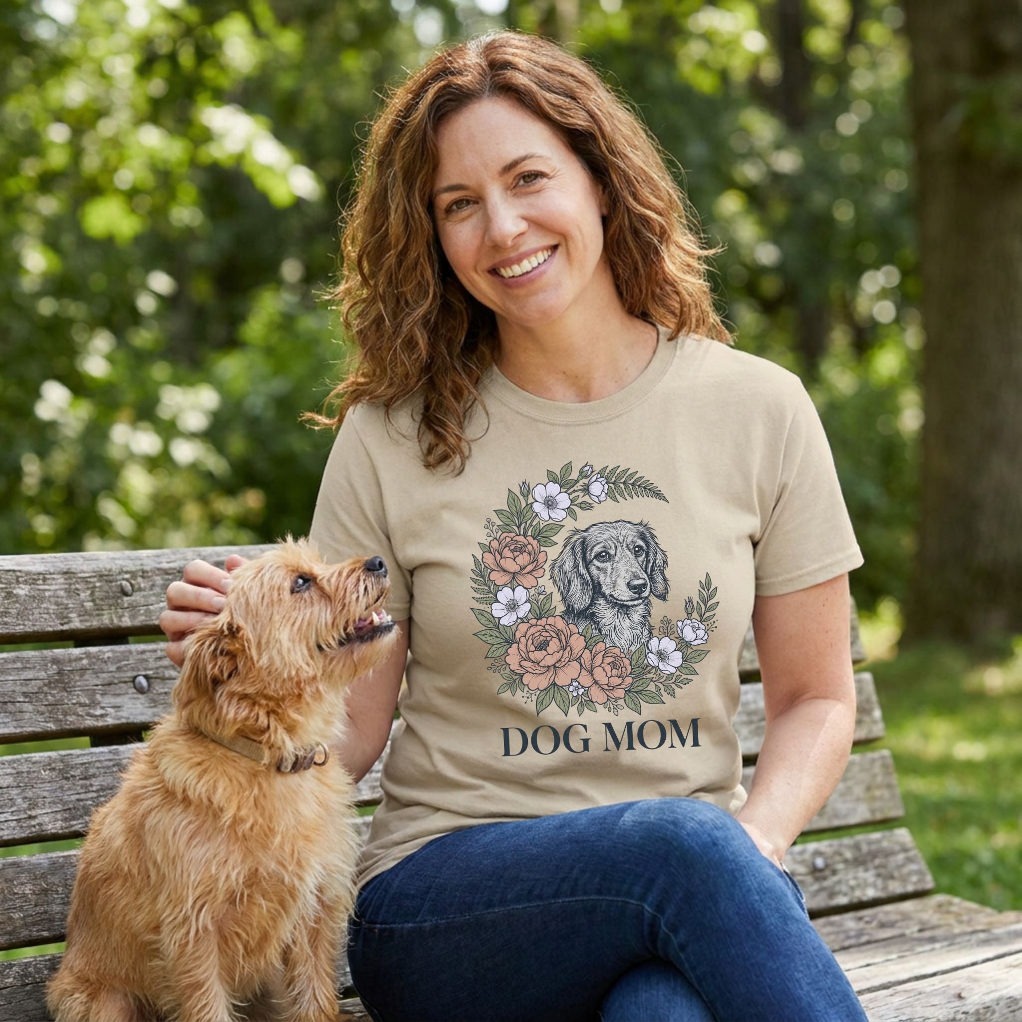 Woman in a tan "Dog Mom" Gildan Softstyle t-shirt with a floral dog portrait design, petting a small dog.