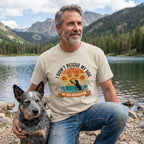 Man in a beige "I Didn't Rescue My Dog, My Dog Rescued Me" tee with a blue heeler by a lake.