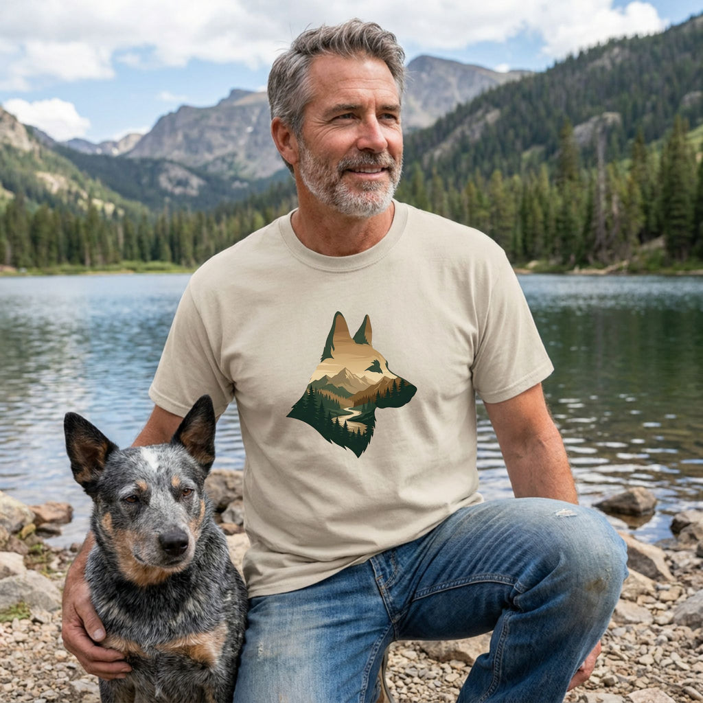 Man in a tan Gildan Softstyle® t-shirt with a mountain silhouette dog design, kneeling with a rescue dog by a lake.