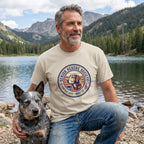 Man in a tan Senior Rescue Society t-shirt with a dog graphic, kneeling by a lake with a blue heeler.