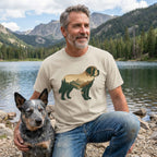 Man in a tan Mountain Dog T-Shirt with a scenic dog graphic, kneeling beside a blue heeler by a lake.