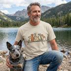 Man in a beige "The Pack" t-shirt with a line of diverse cartoon dogs, kneeling with a blue heeler by a lake.