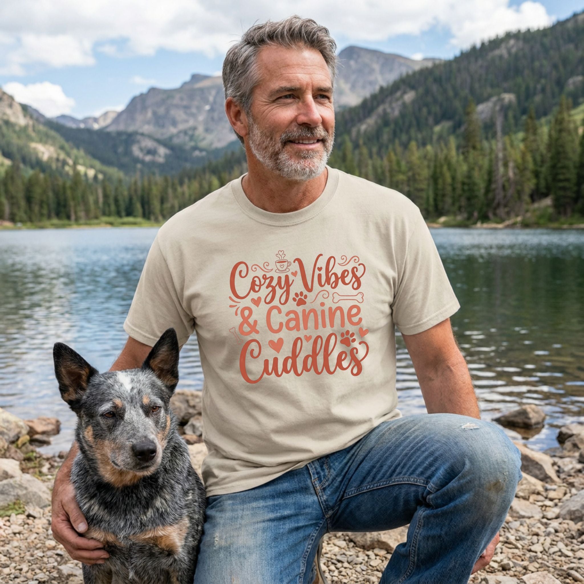 Man in a beige "Cozy Vibes & Canine Cuddles" Gildan tee with a blue heeler dog by a lake.