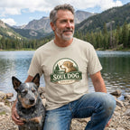 Man in a tan Soul Dog Society logo t-shirt with a blue heeler dog by a lake and mountains.