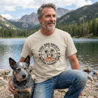 Man in a tan "Soul Dog Society Second Chances Club" tee with a blue heeler dog by a lake.
