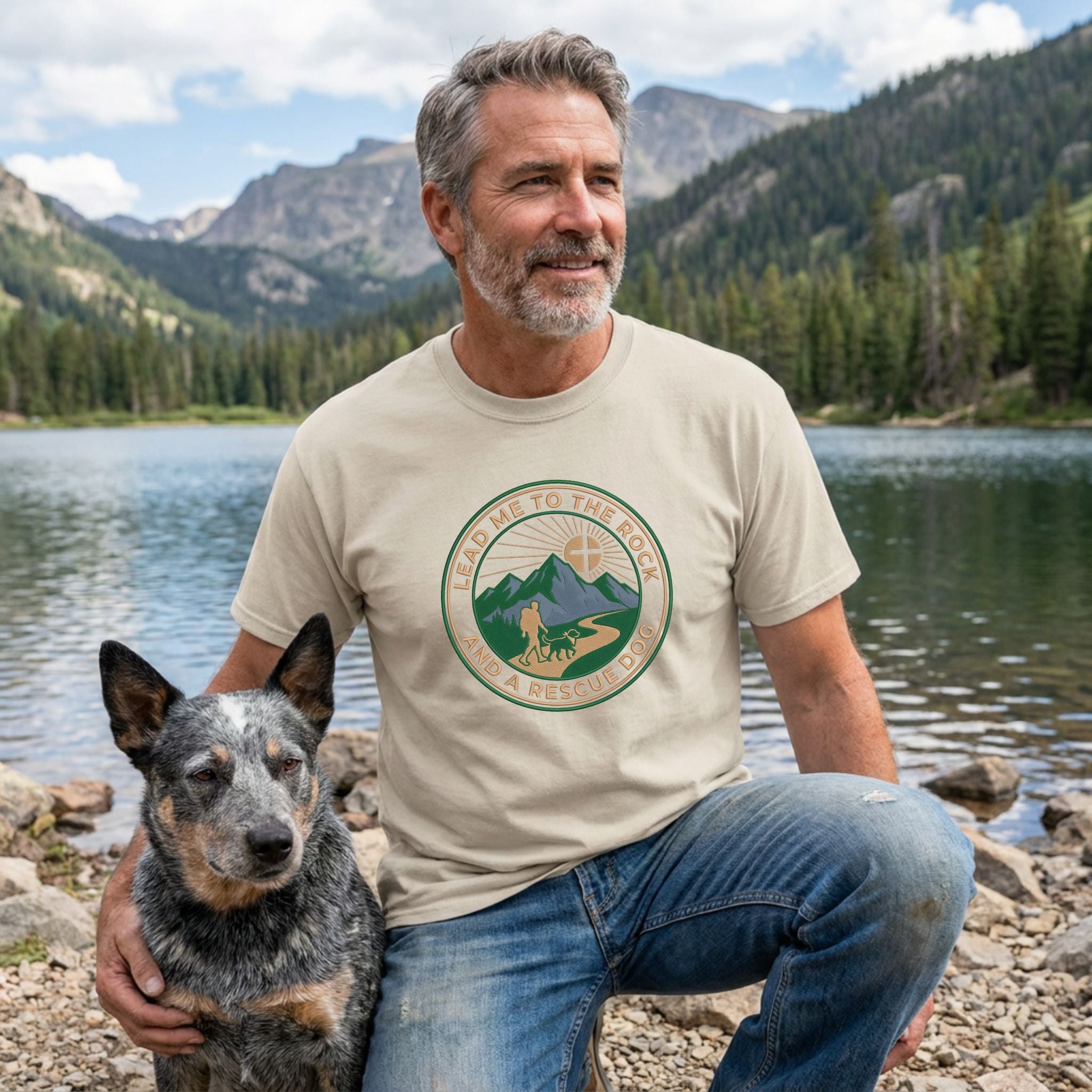 Man in a beige "Lead Me To The Rock" graphic tee with a rescue dog by a mountain lake.