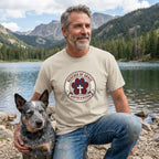 Man in a sand-colored "Rescued By Grace" Gildan Softstyle tee with a blue heeler dog by a lake.