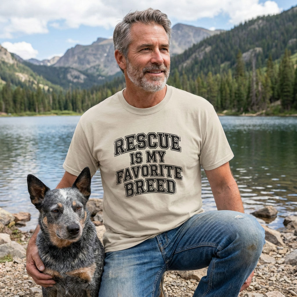 Man in a sand-colored "Rescue Is My Favorite Breed" t-shirt with a blue heeler dog by a lake.