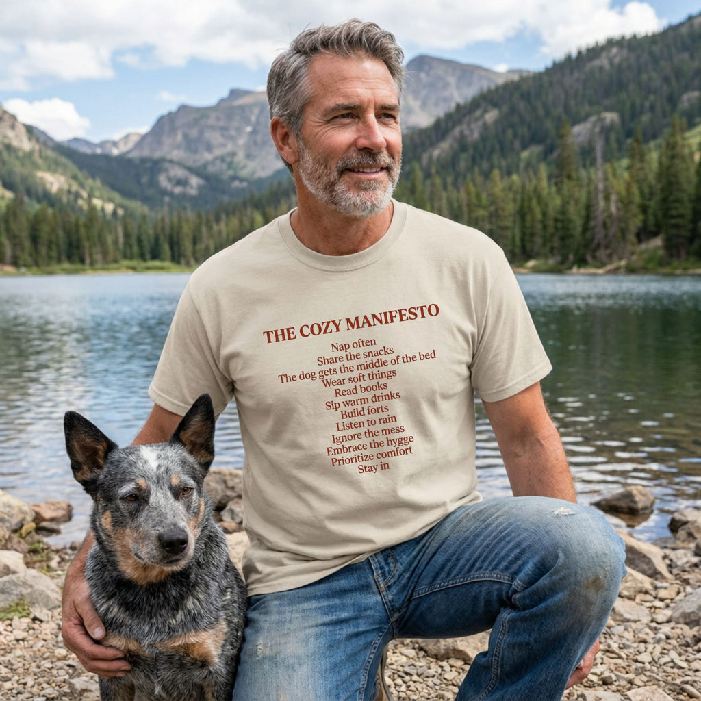 Man in a light beige "The Cozy Manifesto" t-shirt with a rescue dog by a lake.