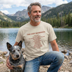 Man in a light beige "The Cozy Manifesto" t-shirt with a rescue dog by a lake.