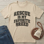 Beige Gildan Softstyle t-shirt with "RESCUE IS MY FAVORITE BREED" in distressed black text, next to jeans and dog treats.