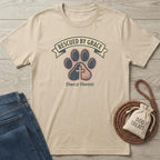 Beige cotton t-shirt with "Rescued By Grace" banner, paw print with cross, and "Paws & Prayers" text.