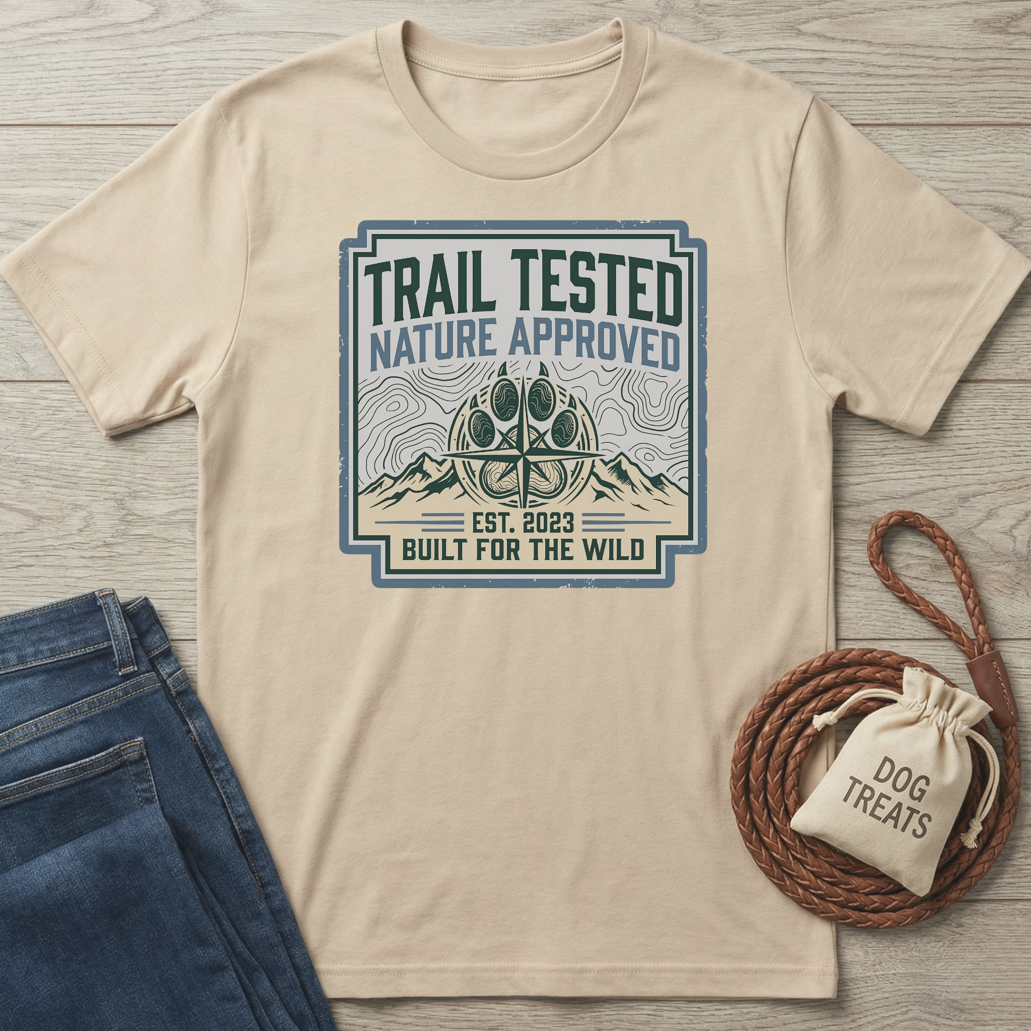 Beige t-shirt with 'Trail Tested, Nature Approved' graphic on a wooden surface with jeans and a dog treat bag.