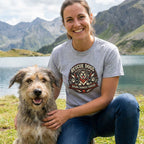 Woman in a grey "Rescue Dogs Motorcycle Club" tee with a dog by a lake and mountains.