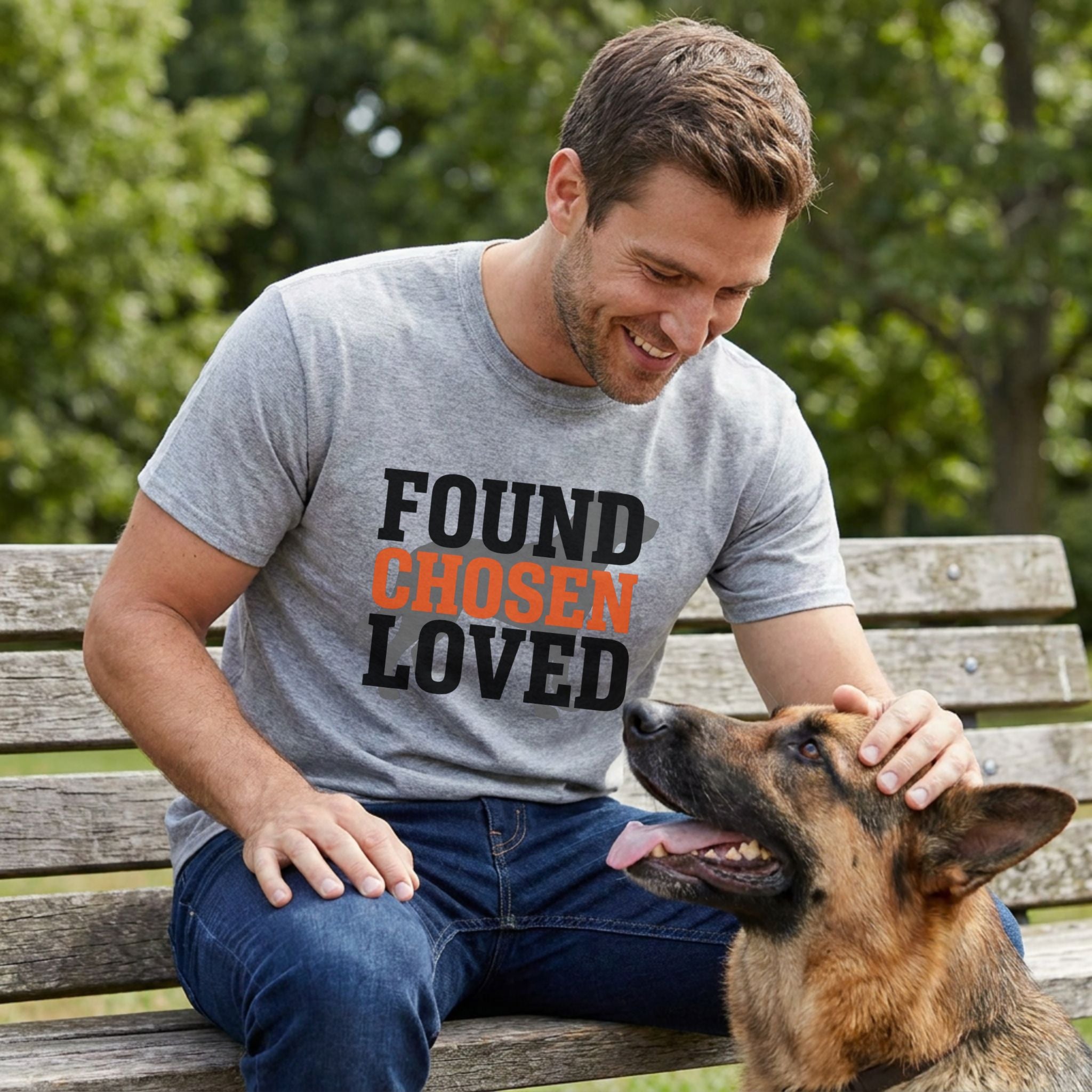 Man in a grey "Found Chosen Loved" t-shirt petting a German Shepherd on a park bench.