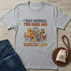 Light grey "Normal 2 Dogs Ago Rescue Life" t-shirt with cartoon dogs, next to jeans and dog treats.
