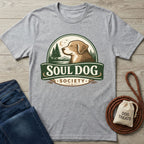 Light grey Soul Dog Society logo t-shirt with a brown dog, trees, and moon design, next to jeans and dog treats.