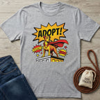 Light grey Gildan Softstyle t-shirt with a comic-style "ADOPT!" superhero dog graphic.