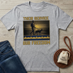 Gray "Their Service Our Freedom" t-shirt with a soldier and dog graphic, displayed on wood with jeans and dog treats.