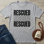 Gray "Rescued" t-shirt with bold black text, folded jeans, braided leash, and "DOG TREATS" bag.