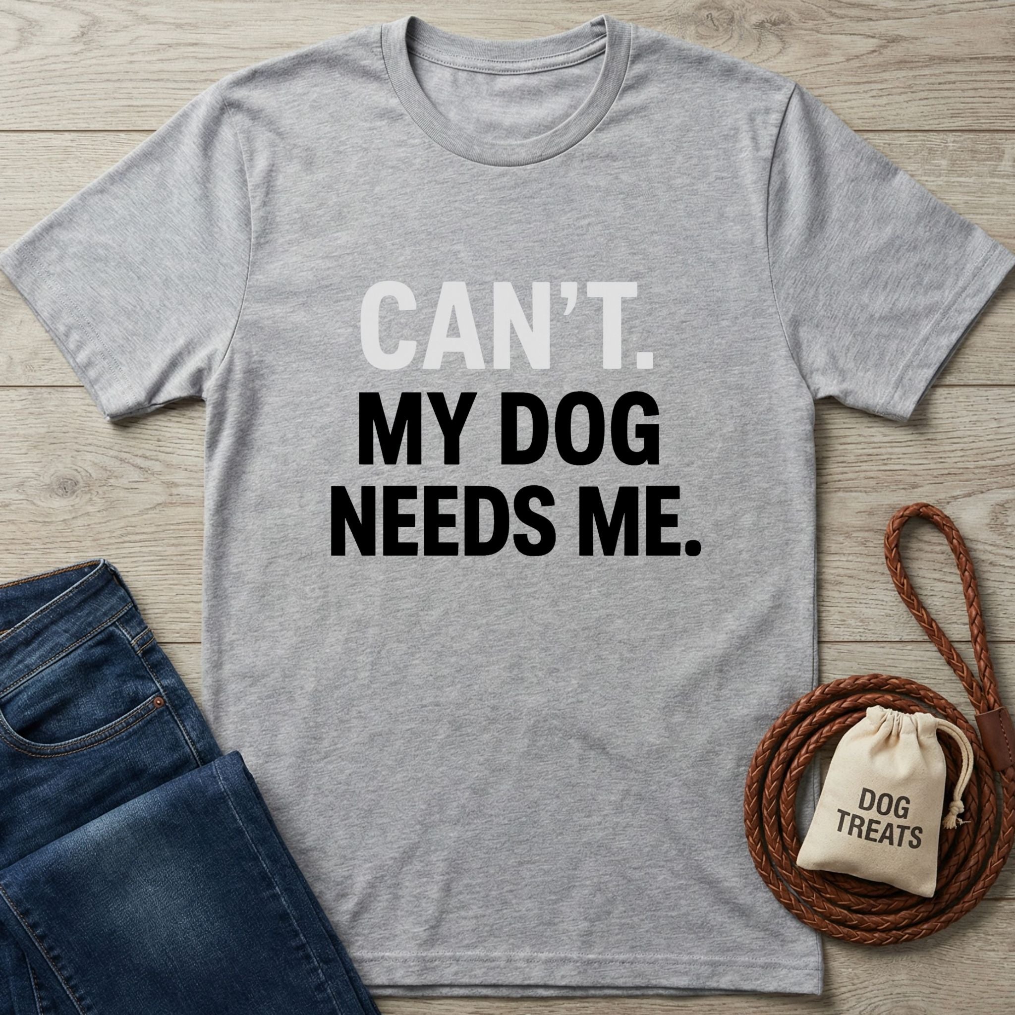 Heather grey Gildan Softstyle t-shirt with "CAN'T. MY DOG NEEDS ME." text, next to jeans and dog treats.