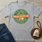 Gray t-shirt with 'Homebody Rescue Club' graphic on a wooden surface with jeans and dog treats bag.