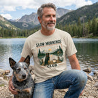 Man wearing a beige Slow Mornings Club graphic t-shirt featuring a dog and lakeside sunrise, kneeling beside his dog.