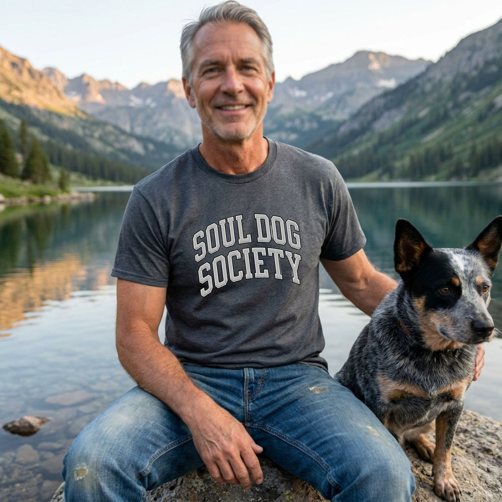 Man in a grey "Soul Dog Society" t-shirt and jeans with a blue heeler dog by a mountain lake.