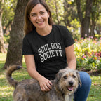 Woman in black "Soul Dog Society" t-shirt, blue jeans, petting a scruffy dog in a park.