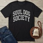 Black cotton t-shirt with "SOUL DOG SOCIETY" in arched silver text, beside blue jeans and dog accessories.
