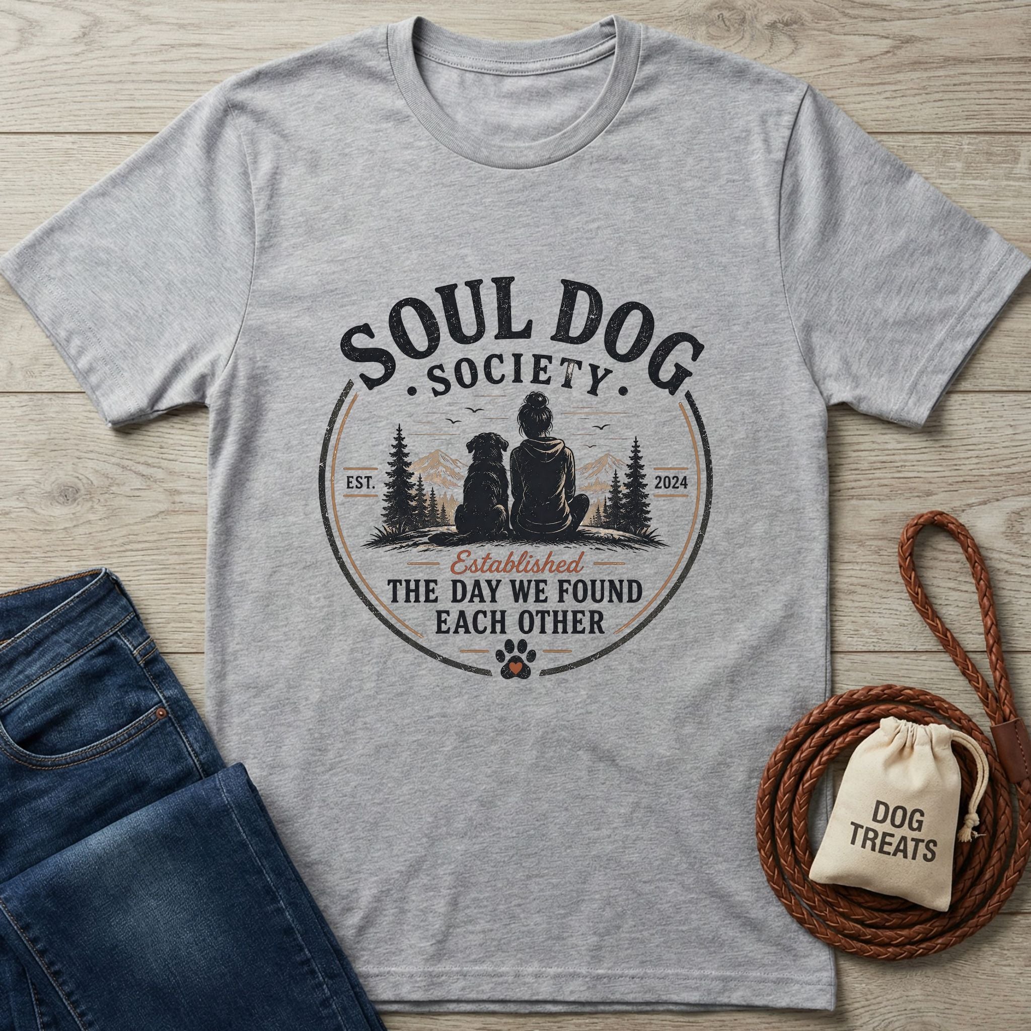 Light grey Gildan Softstyle® t-shirt with "Soul Dog Society" graphic of a person and dog, beside jeans and dog treats.