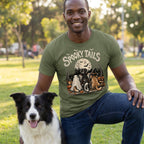 Man wearing an olive green Spooky Tails graphic tee featuring three costumed dogs by a lake under a full moon.