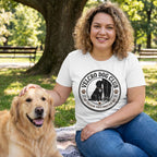 Woman wearing a white crewneck t-shirt with a black Velcro Dog Club graphic, sitting in a park with a golden retriever.