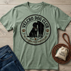 Sage green crewneck t-shirt featuring a Velcro Dog Club graphic with a dog sitting by a person's legs on a wooden background.