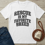 White cotton t-shirt with "RESCUE IS MY FAVORITE BREED" in distressed black text, next to jeans and dog treats.