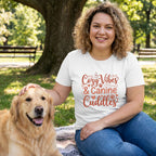 Woman in a white "Cozy Vibes & Canine Cuddles" t-shirt petting a golden retriever in a park.