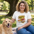 Woman in white "Adopt Dog Comic" tee with a superhero dog graphic, petting a golden retriever in a park.