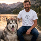 Man in a white Gildan Softstyle Pure Love T-shirt with a dog-heart logo, petting a fluffy Malamute by a lake.