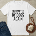 White Gildan Softstyle® t-shirt with "DISTRACTED BY DOGS AGAIN" in black text, next to jeans, a leash, and dog treats.