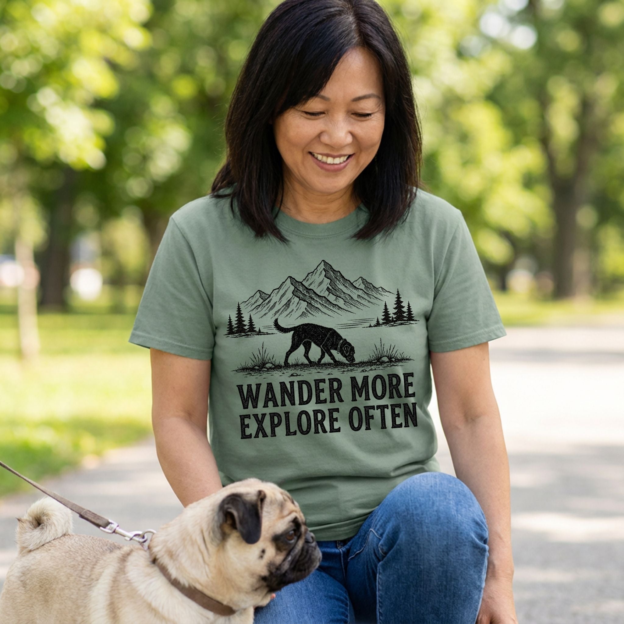 Woman wearing a sage green Wander More T-shirt with a black mountain and dog graphic while walking a pug outdoors.